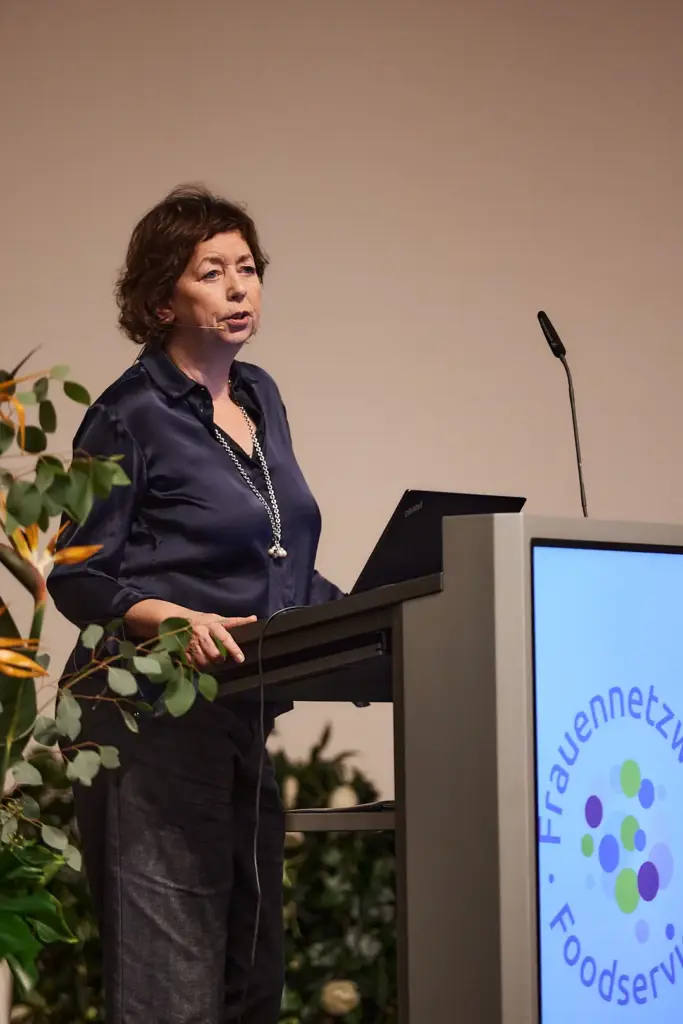 Eine Frau spricht während der Veranstaltung „Frauenforum Foodservice“ mit Laptop und Mikrofon an einem Podium und teilt Neuigkeiten mit, neben ihr sind Pflanzen zu sehen.