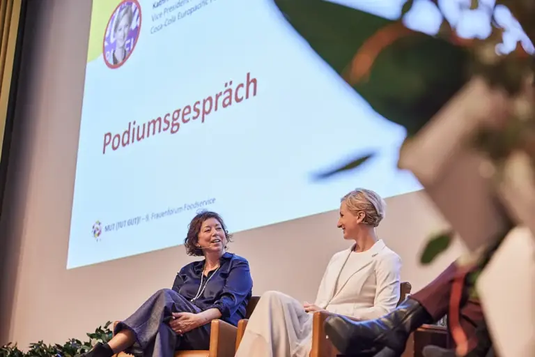 Zwei Frauen sitzen bei einer Podiumsdiskussion auf der Bühne und unterhalten sich. Auf der Leinwand hinter ihnen werden „Podiumsgespräch“ und „Frauenforum Foodservice Neuigkeiten“ angezeigt.