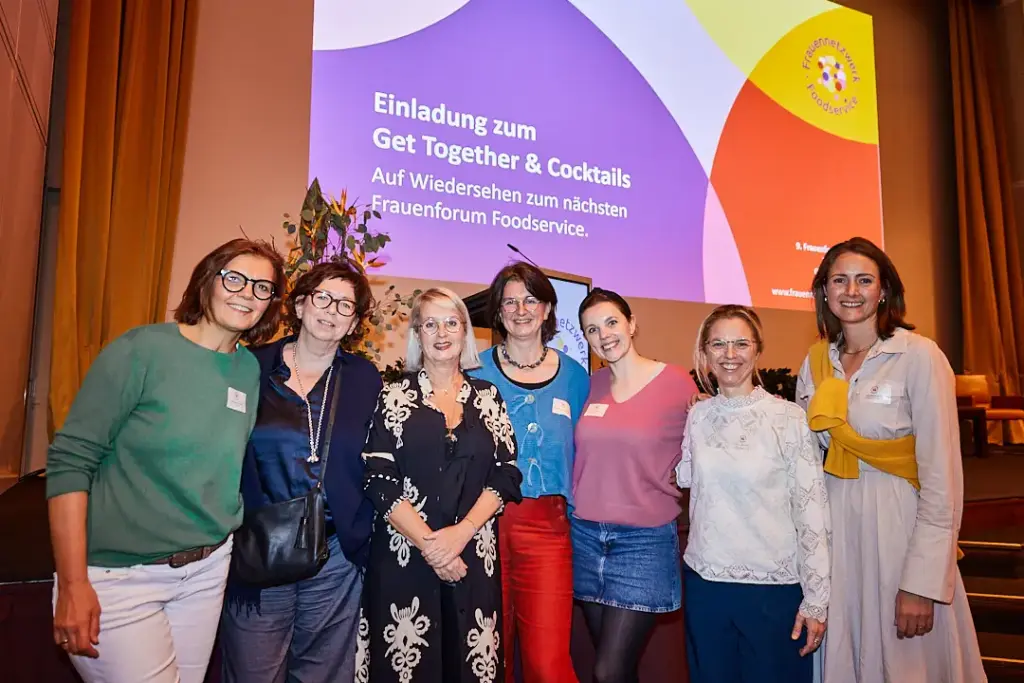 Sieben Frauen stehen lächelnd vor einer farbenfrohen Event-Leinwand bei einem Frauenforum Foodservice-Treffen und feiern die neuesten Frauenforum Foodservice-Neuigkeiten.