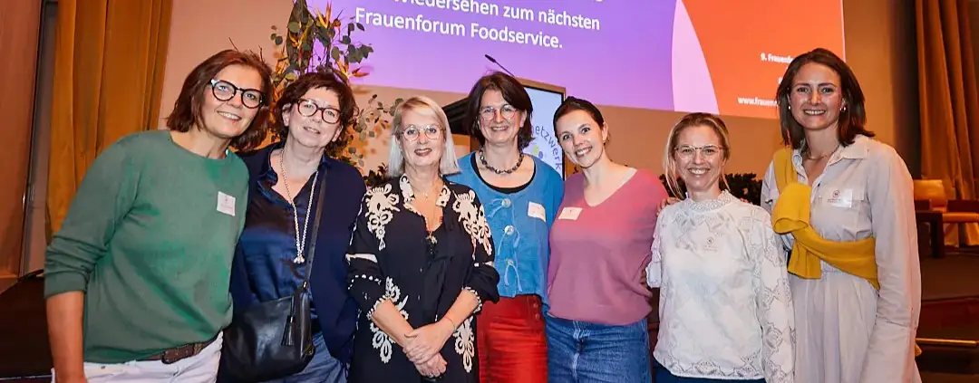 Sieben Frauen stehen lächelnd bei einer Indoor-Veranstaltung des Frauenforum Foodservice zusammen, im Hintergrund läuft ein bunter Bildschirm, auf dem Neuigkeiten verbreitet werden.