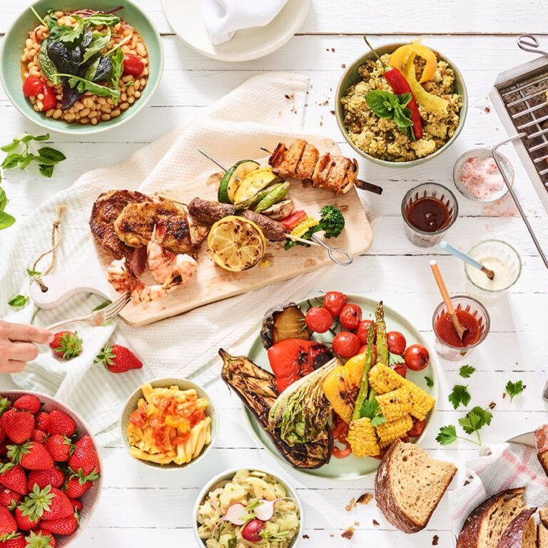 Ein farbenfroher Tisch mit gegrilltem Fleisch, Gemüse, Salaten, Erdbeeren, Brot und Dips auf weißem Holz unterstreicht die Nachhaltigkeit im Catering mit frischen, regionalen Zutaten.