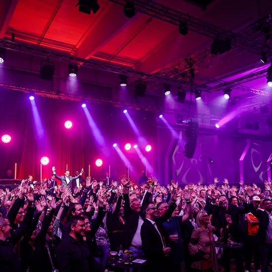 Große Menschenmenge bei einem Indoor-Konzert mit violettem Bühnenlicht und Menschen, die ihre Hände heben.