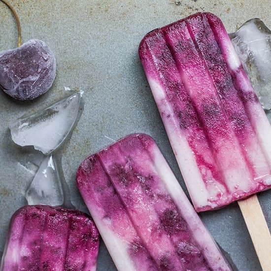 Drei Kirsch-Eis am Stiel mit Eiswürfeln und einer gefrorenen Kirsche auf grauem Untergrund – ein idealer Leckerbissen.