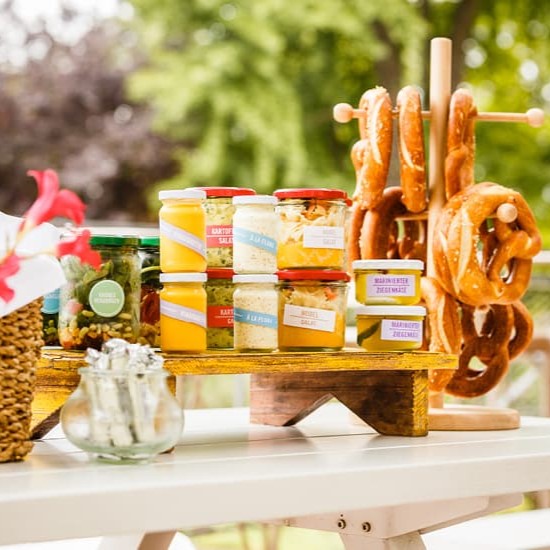 Gläser mit Brotaufstrichen und Pickles sowie frischen Brezeln auf einem Ständer, präsentiert auf einem Tisch im Freien.