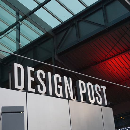 Großes weißes DESIGN POST-Schild an einem modernen Gebäude mit architektonischen Elementen aus Glas und Stahl darüber.