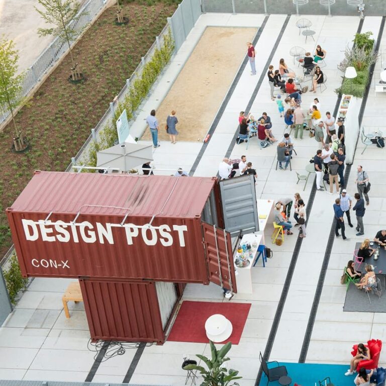 Urbane Sommerparty - Luftaufnahme von Menschen, die sich in einem städtischen Außenbereich an einem Schiffscontainer mit der Aufschrift „DESIGN POST“ versammeln.