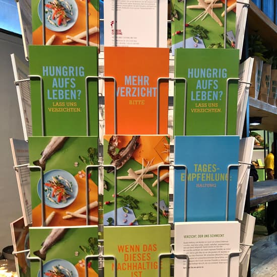 In einem Metallständer werden farbenfrohe Broschüren mit deutschem Text ausgestellt, die für einen nachhaltigen Lebensstil werben und das Thema Nachhaltigkeit im Catering hervorheben.