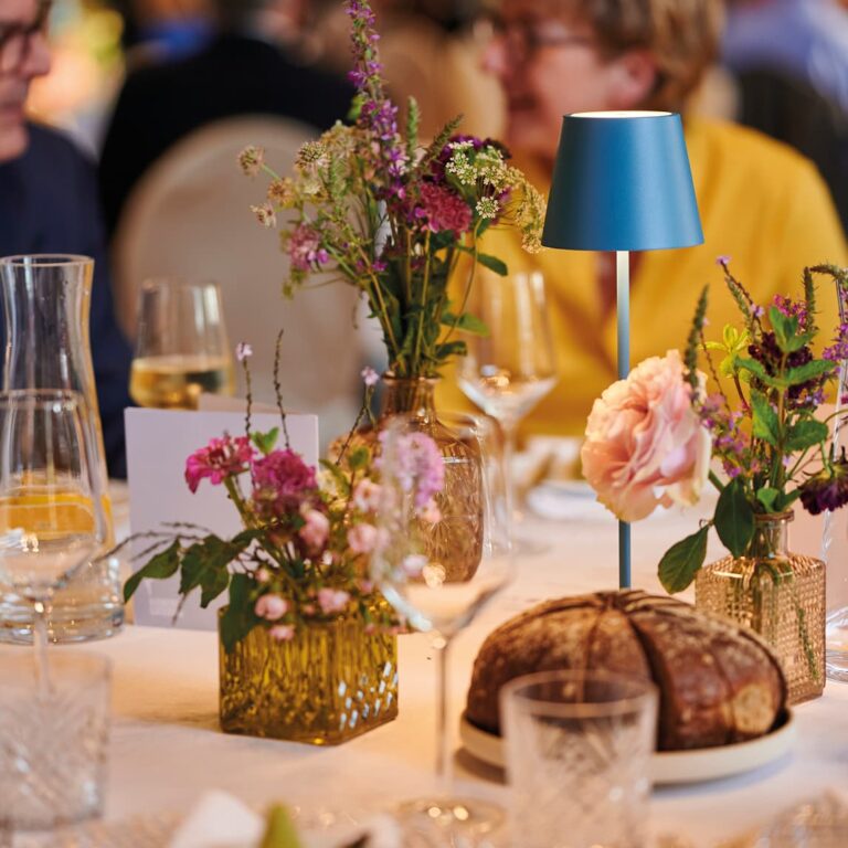 Gedeckter Tisch mit Blumen, Brot, Weingläsern und einer blauen Lampe; im Hintergrund unterhalten sich Menschen.