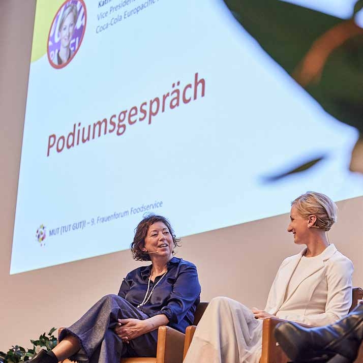 Zwei Frauen sitzen auf der Bühne vor einem Bildschirm mit der Aufschrift „Podiumsgespräch“ und unterhalten sich bei einer Veranstaltung über Themen wie Nachhaltigkeit im Catering.