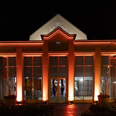 Ein Gebäude mit hohen Fenstern, die nachts orangefarbenes Licht spenden und durch dessen Eingang man Menschen sehen kann.