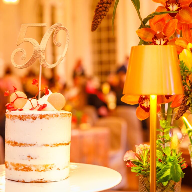 Eine weiße Torte mit einem 50-Dollar-Topper steht neben orangefarbenen Blumen und einer goldenen Lampe bei einer Feier.