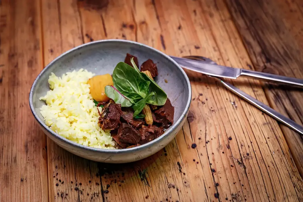 Eine Schüssel mit gelbem Reis, Rindfleischeintopf, Gemüse und einer Kartoffel auf einem Holztisch, daneben Besteck.