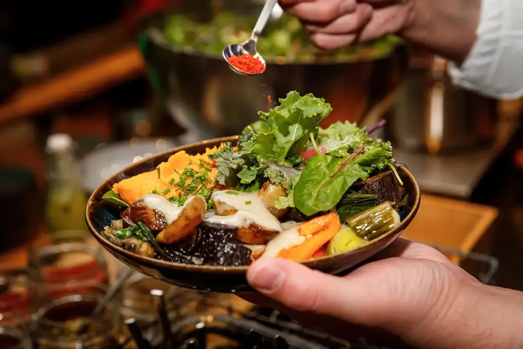 Eine Hand hält eine bunte Gemüseschüssel, während eine andere Hand rote Gewürze darüber streut.