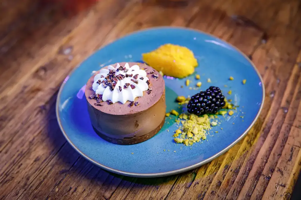Schokoladenmousse-Dessert mit Schlagsahne, Orangenscheibe, Brombeere und zerbröselten Pistazien auf einem blauen Teller.
