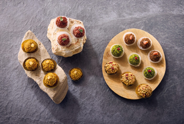 Verschiedene bunte Trüffel, angeordnet auf Stein- und Holzbrettern auf einer grau strukturierten Oberfläche.