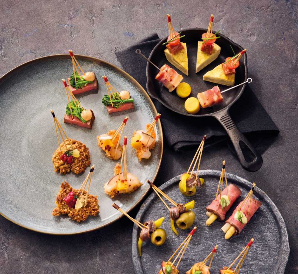 Verschiedene mundgerechte Tapas mit Zahnstochern auf Tellern und einer Bratpfanne, serviert auf einer dunkel strukturierten Oberfläche.