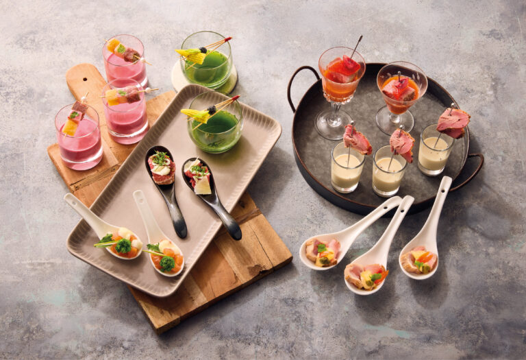 Verschiedene Fingerfoods, darunter Suppen im Glas, Spieße und Löffelgerichte, kunstvoll dekoriert auf Holz-, Metall- und Porzellantabletts.