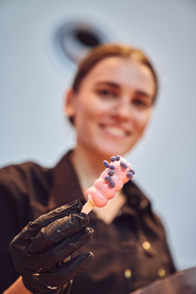 Patissière von Kirberg Catering hält ein rosafarbenes, garniertes Popsicle in der Hand. Nahaufnahme.