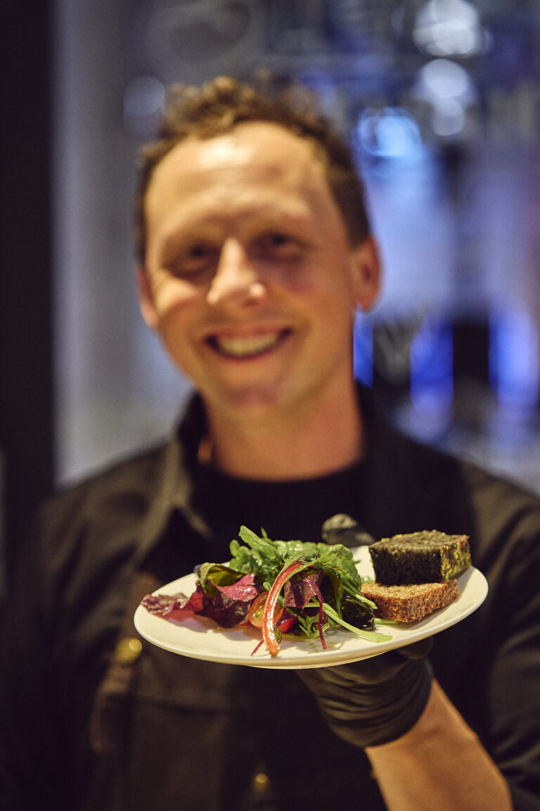 Lächelnder Kirberg Catering Mitarbeiter zeigt ein Salat-Menü für die #wtf24 in der Grand Hall Zollverein.