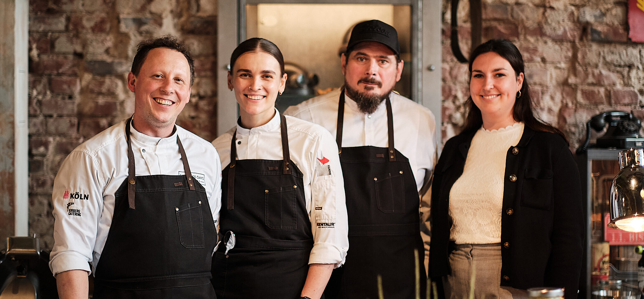 Vier Personen, drei in Kochuniformen und eine in Freizeitkleidung, stehen lächelnd in einer rustikalen Küche.