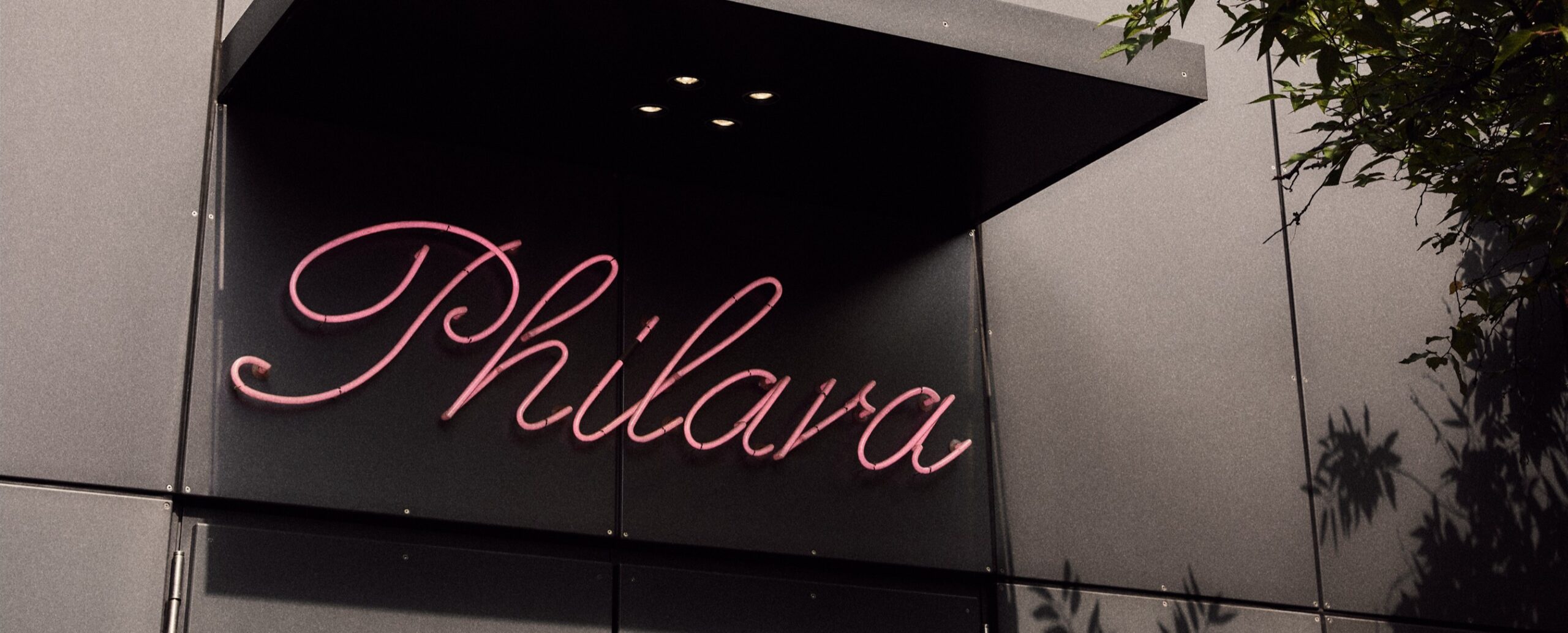 Rosa Leuchtreklame mit der Aufschrift Philara auf einem modernen grauen Gebäude, mit Baumschatten auf der rechten Seite.