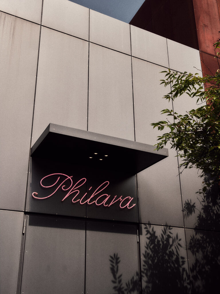 Ein rosafarbenes Neonschild mit der Aufschrift Philara an einem modernen grauen Gebäude, mit Baumschatten an der Wand, spiegelt das Engagement des Lokals für Nachhaltigkeit im Catering wider.
