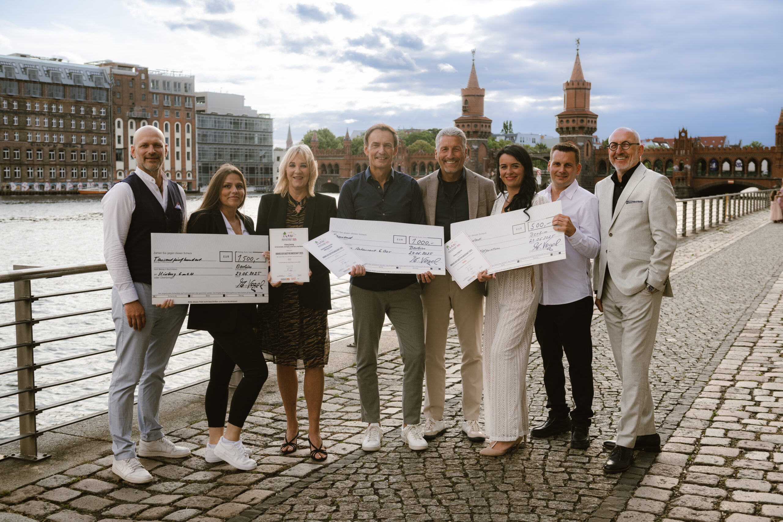 Acht Personen stehen im Freien an einem Fluss, halten große Schecks in der Hand und lächeln in die Kamera.