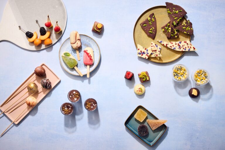 Verschiedene Desserts: Schokoladenrinde, Trüffel, Kuchen und Obstspieße auf pastellfarbenen Tellern und Tabletts angeordnet.