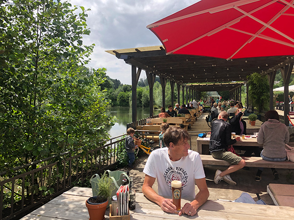 Ein Mann mit Bier sitzt an einem sonnigen Tag in einem Café am Flussufer, wo Menschen unter großen roten Sonnenschirmen speisen.