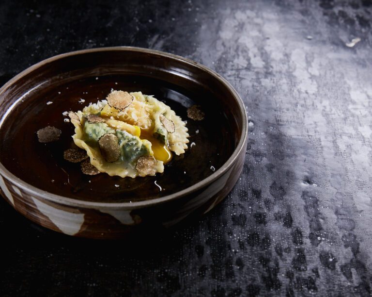 Ein Gourmet-Ravioli-Teller mit gehobeltem Trüffel auf einem braunen Keramikteller, der auf einer dunklen, strukturierten Oberfläche steht.