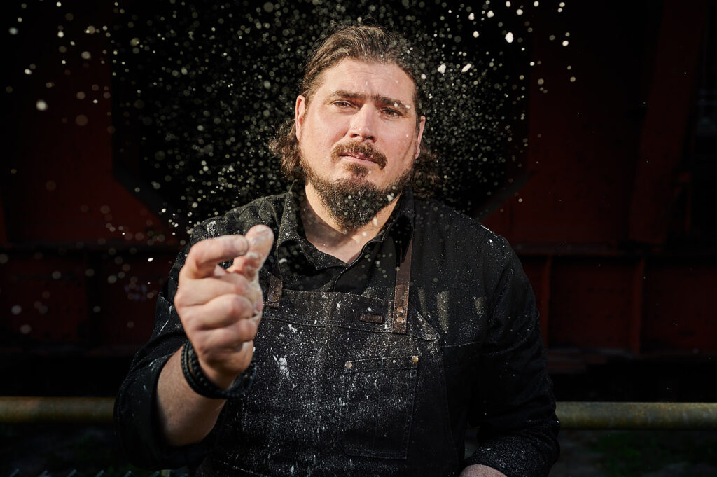 Volker Beuchert in einer schwarzen Schürze streut Mehl, wobei um ihn herum Mehlstaub in der Luft schwebt.