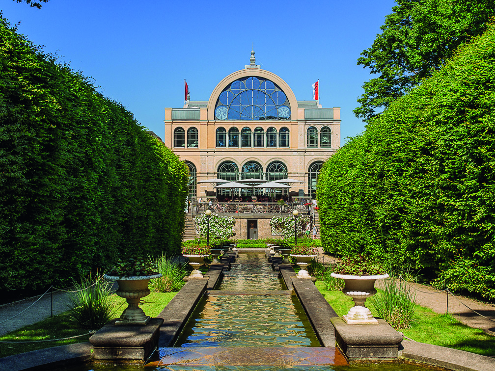 Großes gläsernes Gebäude mit Bogenfenstern, formalen Gärten, Heckenmauern und einem zentralen reflektierenden Becken.