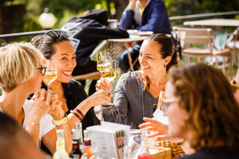 Vier Frauen, die lächelnd und mit einem Glas Weißwein an einem Cafétisch im Freien in der Sonne anstoßen.