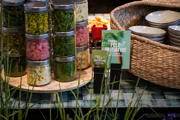 Gläser mit eingelegtem Gemüse und Kräutern sind neben gestapelten Schalen und einem geflochtenen Korb ausgestellt.