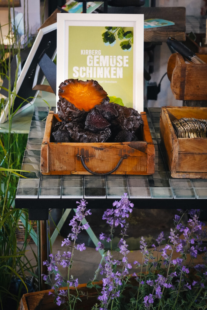 Ein Holztablett mit dunklem Trockengemüse steht auf einem gekachelten Tisch, im Hintergrund stehen Blumen und ein Schild.