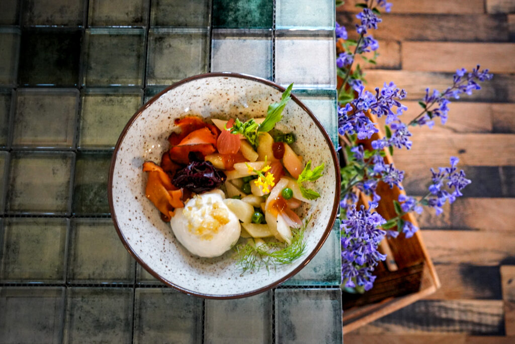 Eine Schale mit buntem Gemüse und Käse auf einem gekachelten Tisch, daneben lila Blumen.