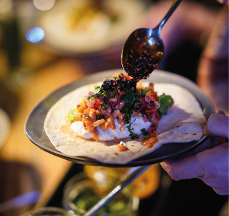 Eine Hand bedeckt mit einem Löffel eine Tortilla mit Salsa und Kräutern und bereitet einen frischen Taco auf einem Teller zu - ideal für Eventcatering, das auf regionale Produkte setzt.