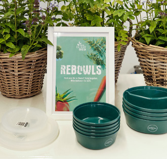 Gestapelte grüne Schalen, ein weiß gerahmtes Rebowls-Schild, Deckel und Körbe mit Blattpflanzen im Hintergrund präsentieren Regionale Produkte im Eventcatering für eine frische und nachhaltige Eventatmosphäre.