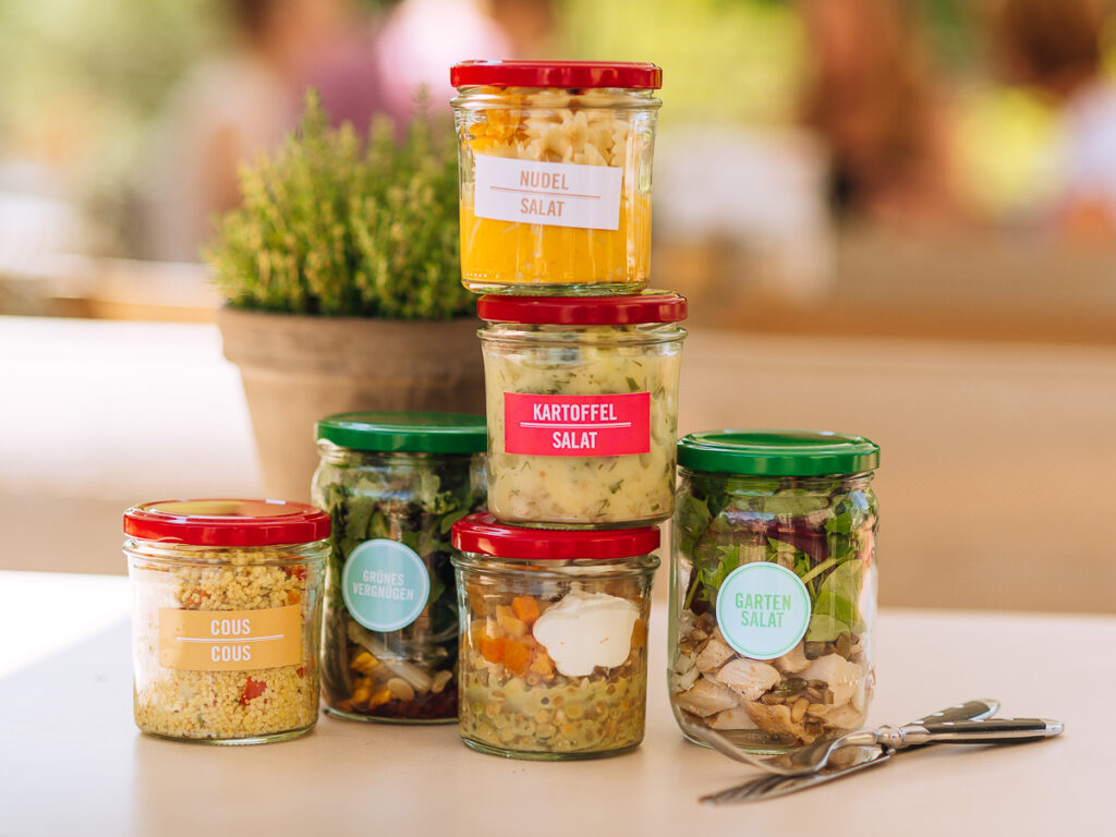 Sechs verschiedene Salatgläser mit bunten Etiketten stapeln sich auf einem Tisch neben Utensilien und einer Topfpflanze.