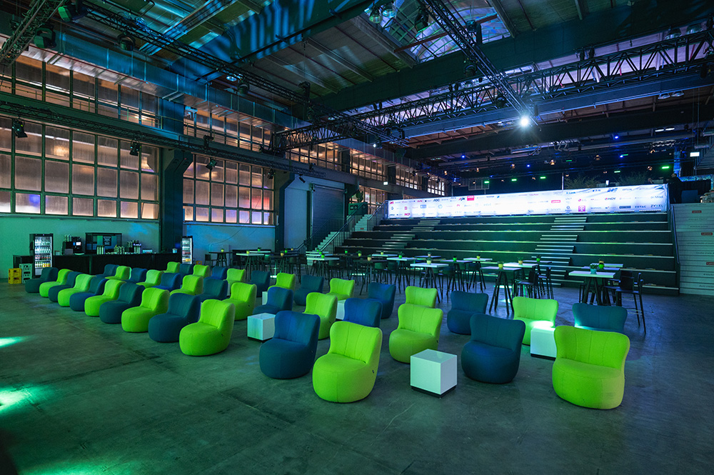 Geräumiger, moderner Veranstaltungssaal mit bunten Loungesesseln, Stehtischen und einer beleuchteten Bühne im Hintergrund.