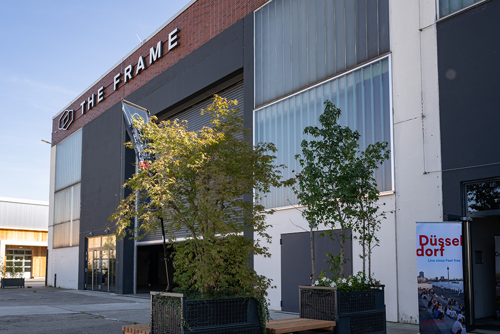 Außenansicht des The Frame-Gebäudes mit Bäumen und einem Düsseldorfer Veranstaltungsplakat am Eingang.