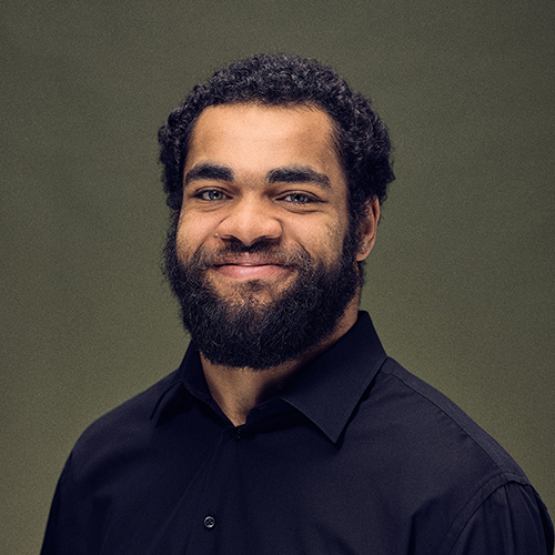 Lächelnder Mann mit Bart und lockigem Haar, der ein schwarzes Hemd trägt, vor einem olivgrünen Hintergrund.