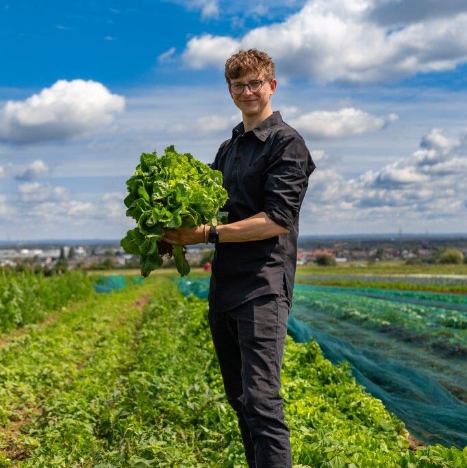 Lächelnde Person in schwarzer Kleidung, die einen großen Salatkopf in einem grünen Feld unter einem blauen Himmel mit Wolken hält.