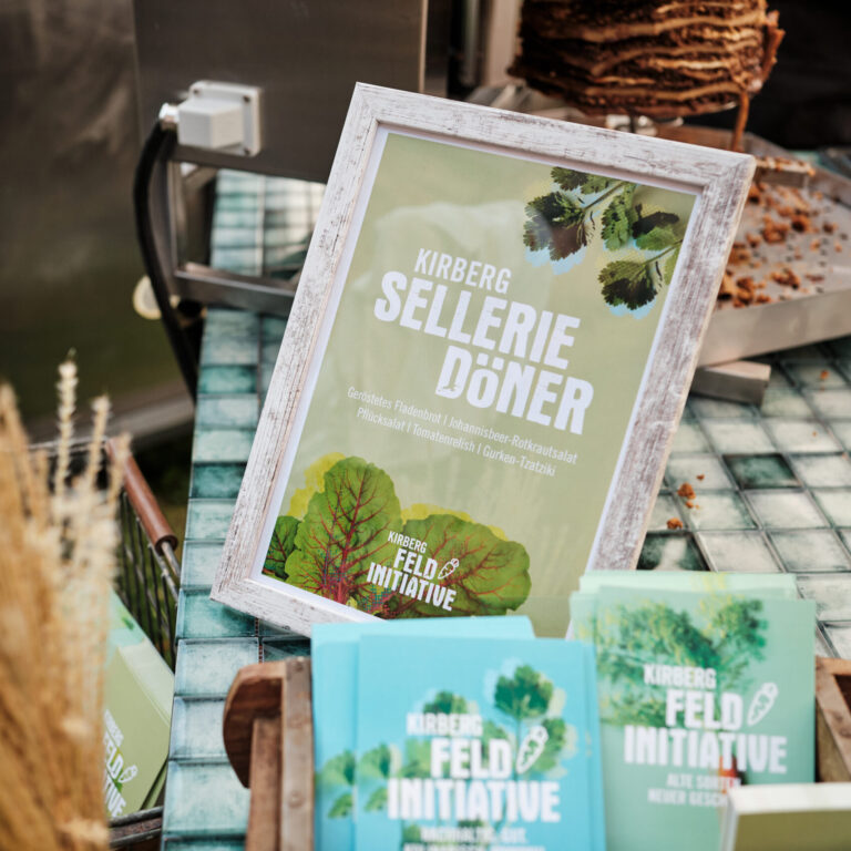Gerahmtes Schild mit der Aufschrift Sellerie Döner auf einem grün karierten Tisch mit Broschüren und Speisen im Hintergrund.
