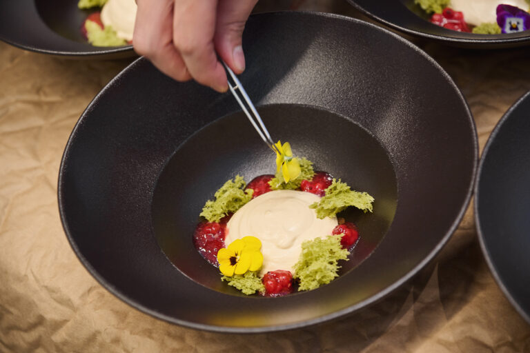 Eine Hand setzt mit einer Pinzette essbare Blumen auf ein Dessert mit Sahne, Beeren und grünem Biskuit.
