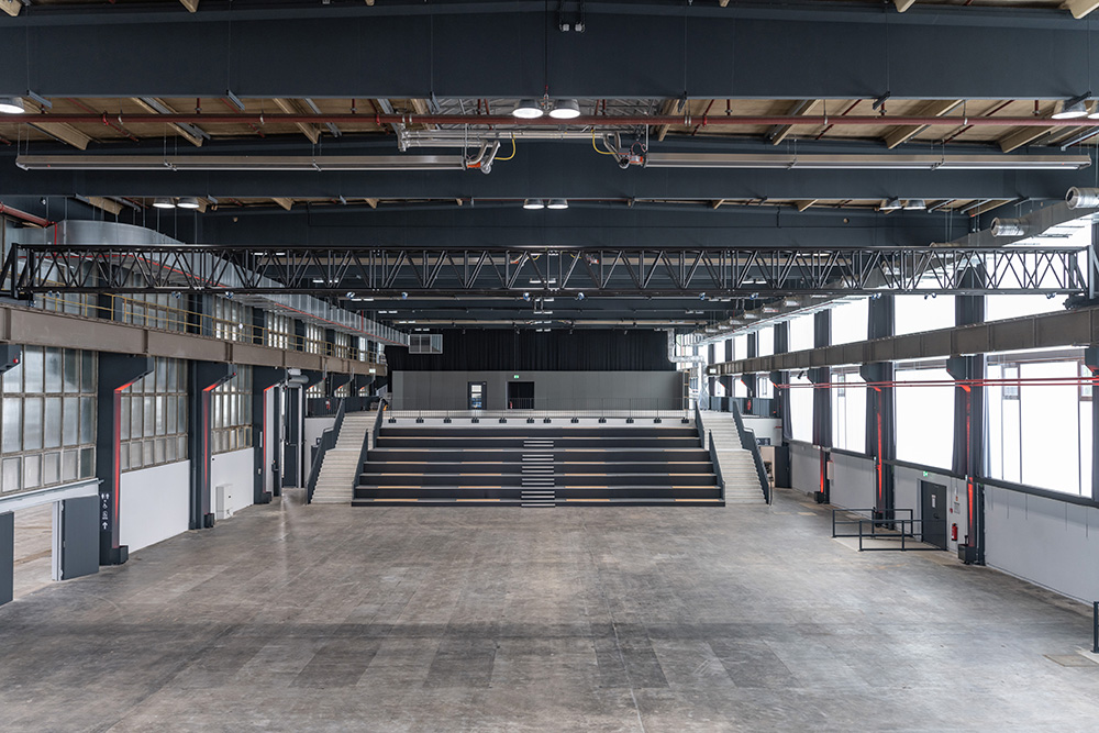 Geräumige leere Industriehalle mit Tribünenplätzen und großen Fenstern an den Wänden.