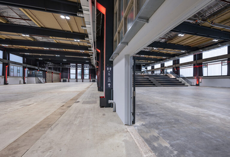 Großzügige, moderne Industriehalle im The Frame in Köln für Events, mit Betonböden, hohen Decken und schwarzen Sitzreihen auf der rechten Seite.