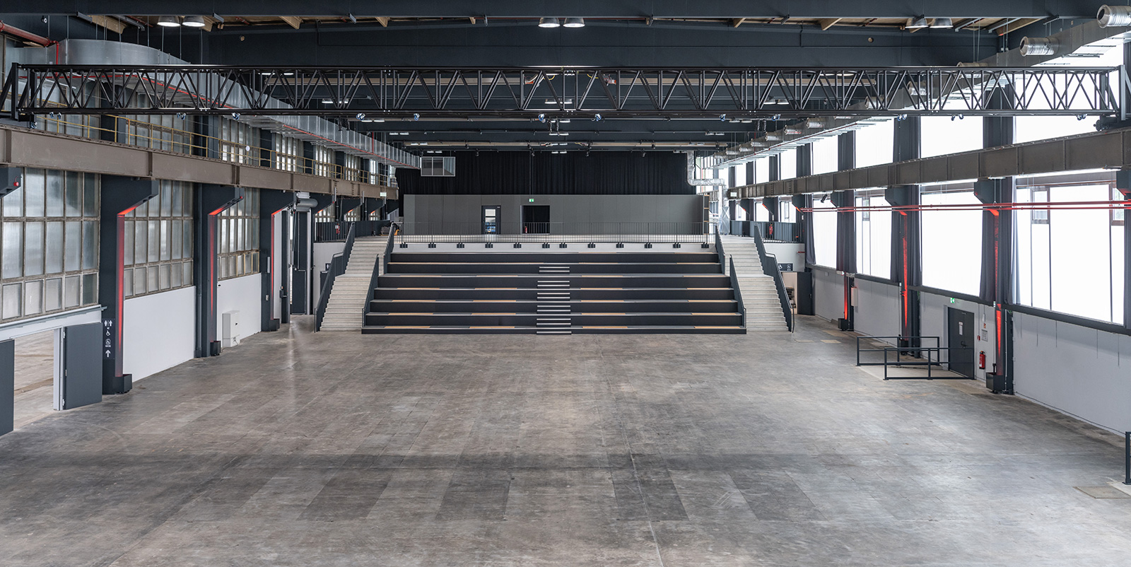 Geräumige, leere Industriehalle mit abgestuften Sitzplätzen und großen Fenstern an den Seiten.