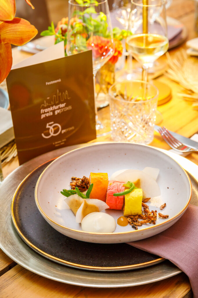 Buntes Gourmet-Dessert mit Fruchtwürfeln, Garnierungen und Soße auf einem gedeckten Esstisch mit Gläsern.