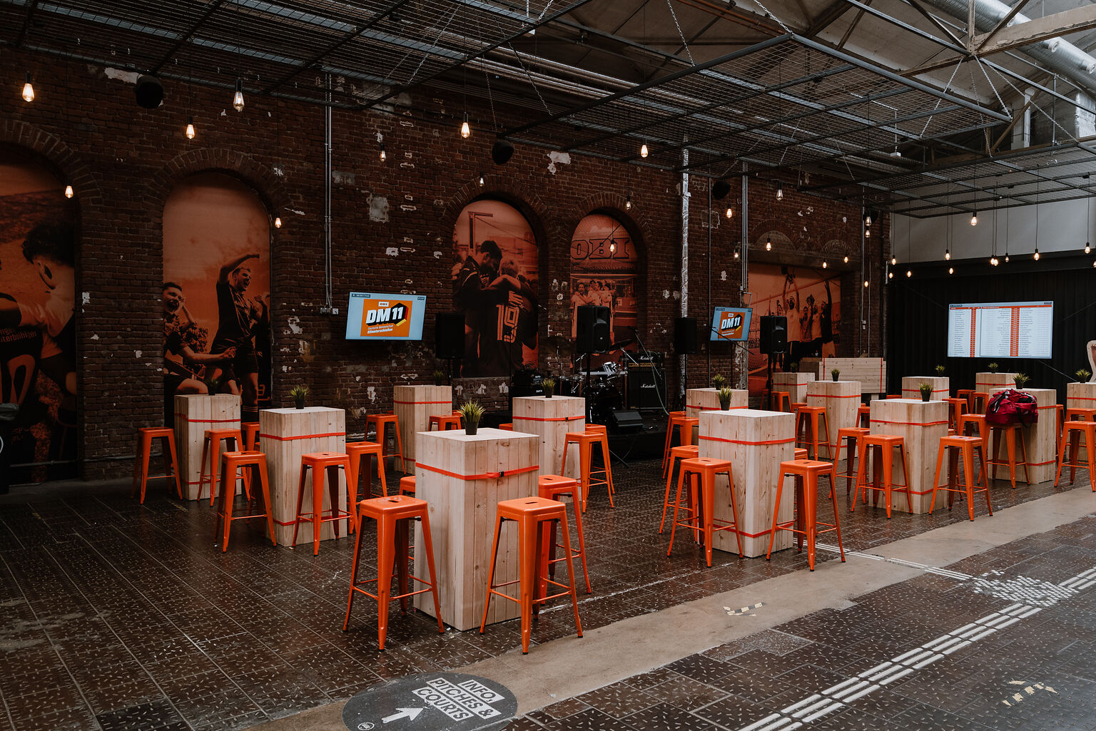 Veranstaltungsraum im Industriestil mit Holztischen, orangefarbenen Hockern, Lichterketten und sportlicher Dekoration - ideal für Catering im BASE Köln - Eventlocation für Business, Kultur & private Feiern.